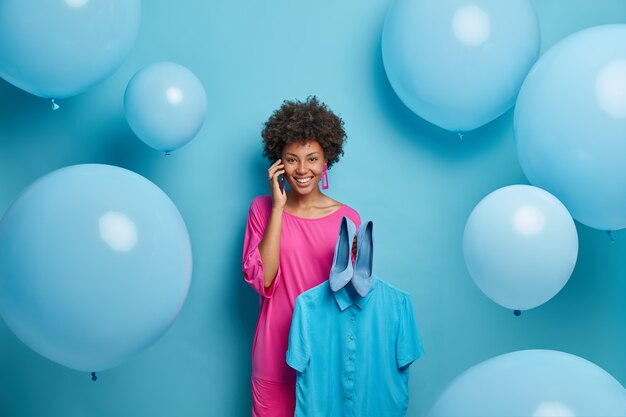 Gorgeous joyful dark skinned woman enjoys telephone conversation with best friend, tries new high heel shoes and shirt on hanger, prepares for bachelorette party, poses against blue wall