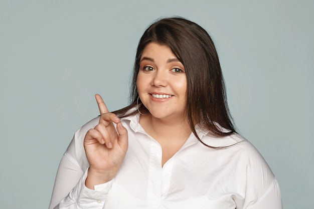 Gorgeous joyful young plus size businesswoman in good mood posing isolated at gray wall, looking with happy confident smile, showing something to you, pointing index finger sideways