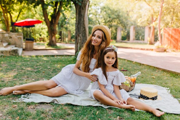 밀짚 모자와 흰색 드레스에 화려한 긴 머리 여자는 좋은 여름날에 딸과 함께 피크닉을 가지고 있습니다. 공원에서 어머니와 함께 시간을 보내는 예쁜 소녀의 야외 초상화.