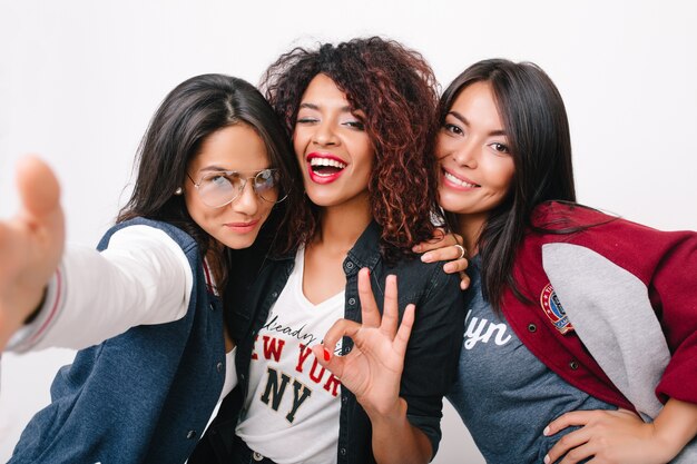 Gorgeous mulatto girl posing with ok sign between latin and asian friends. Indoor portrait of pleased young women from different countries standing together with smiles.