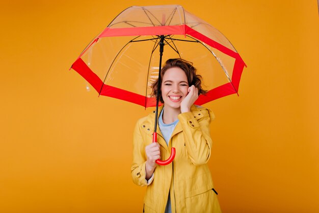 Великолепная бледная девушка в осеннем пальто улыбается с закрытыми глазами под зонтиком. Студийный портрет стильной кавказской женщины с волнистыми волосами, держащей красный зонтик.