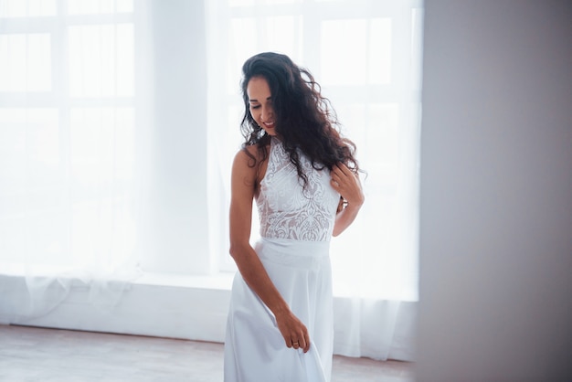 Gorgeous portrait. Beautiful woman in white dress stands in white room with daylight through the windows