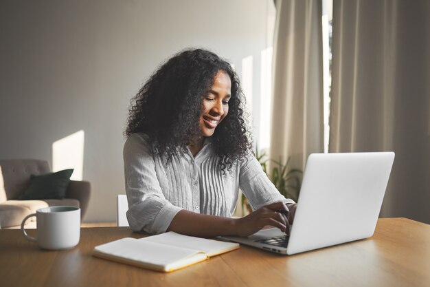 화려한 긍정적 인 젊은 어두운 피부의 여성 블로거가 일반 노트북에서 키보드를 사용하고, 웃고, 그녀의 여행 블로그를위한 새로운 콘텐츠를 만드는 동안 영감을 받고, 일기와 머그잔으로 책상에 앉아