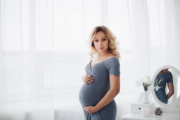 Gorgeous pregnant woman wearing in long gray dress posing at stylish studio embracing her stomach and smiling at camera Pretty blonde mom expacting child Sweet interior of studio