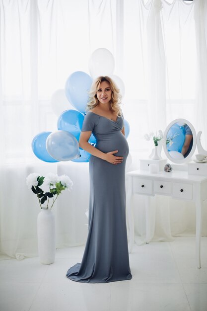 Gorgeous pregnant woman wearing in long gray dress posing at stylish studio embracing her stomach and smiling at camera Pretty blonde mom expacting child Sweet interior of studio