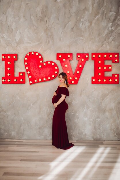 Gorgeous pregnant woman wearing in marsala dress posing touching her belly Beautiful future mother standing on steps of white house Concept of maternity and motherhood