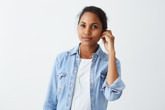 Gorgeous pretty African-American woman with hair bun, dark eyes dressed in white T-shirt, blue jacket, wearing earphones on, listening music, using music app on her mobile phone.