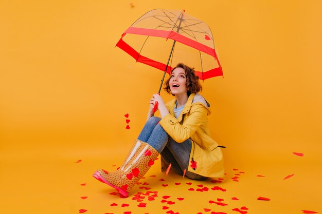 Gorgeous short-haired girl with beautiful eyes posing under parasol. Indoor photo of romantic white female model sitting on yellow floor with umbrella.