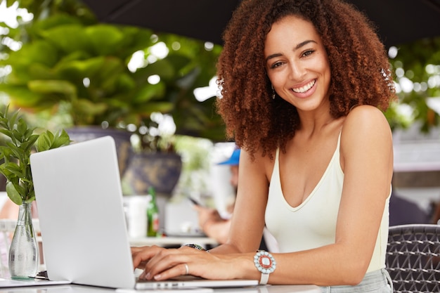 Foto gratuita splendida donna sorridente gode di ricreazione nella caffetteria, ha videochiamate tramite laptop portatile, utilizza l'applicazione.