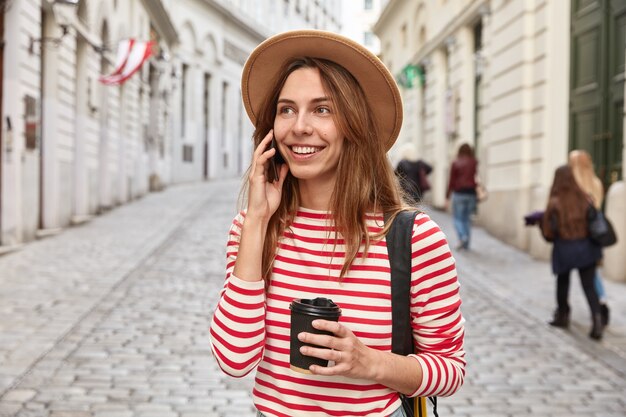 ゴージャスな笑顔の観光客が街を散歩し、電話で会話し、持ち帰り用のコーヒーを飲み、遠くに集中します