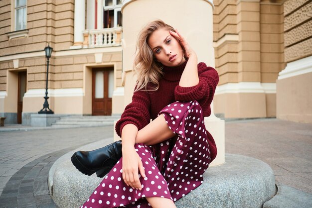 Gorgeous stylish blond girl in knitted sweater confidently looking in camera while posing outdoor