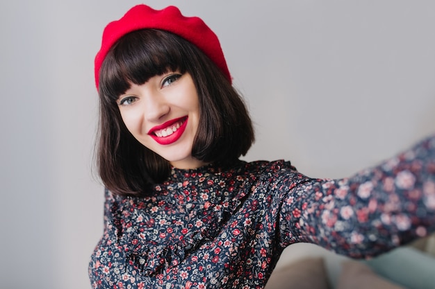 Splendida ragazza bruna alla moda che indossa un abito retrò con stampa floreale che sorride ampiamente mentre fa selfie. ritratto del primo piano di adorabile giovane donna francese in berretto rosso alla moda con l'espressione del viso felice