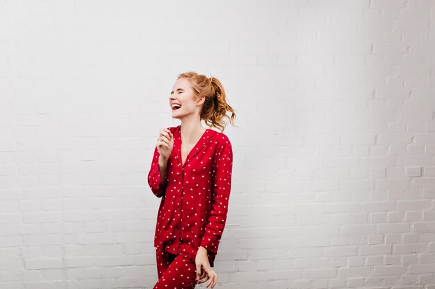 目を閉じて笑っているかわいいパジャマのゴージャスな白人の女の子。朝楽しんでいる赤いパジャマの驚くべき白人のブロンドの女性。