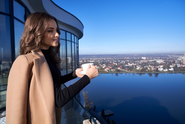 Великолепная женщина пьет кофе, стоя на балконе