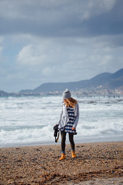 Великолепная женщина в сером балахоне и шляпе стоя и держа камерун пляж в дневное время с морем