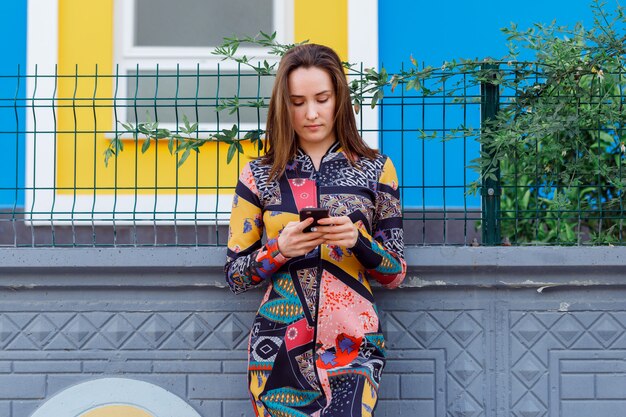 Gorgeous woman leaning on wall and holding a phone in colorful dress in street