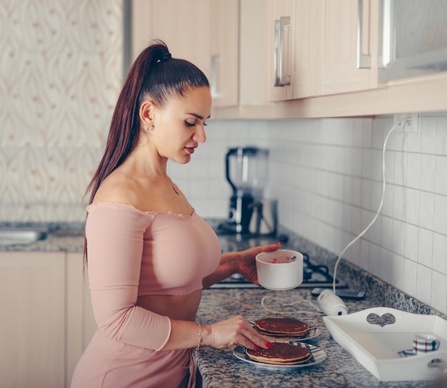 Великолепная женщина поливает смешанную клубнику на блины на кухне в лососево-розовом топе и брюках