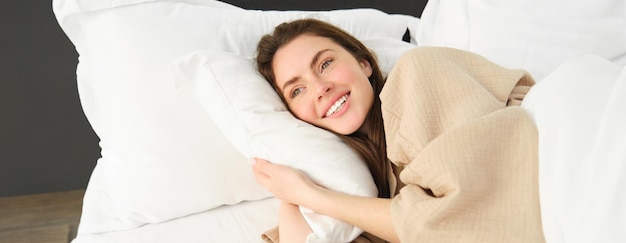 Free photo gorgeous woman resting in hotel room sleeping in bed lying on pillow with eyes opened and smiling