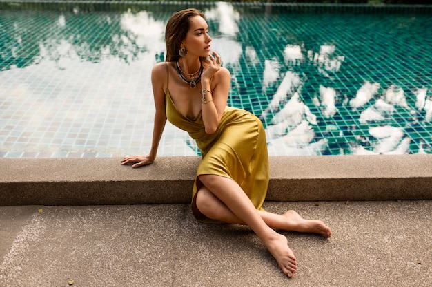 Gorgeous woman in silk dress chilling near pool during tropical vacation. Golden jewelry. Palm trees on background.