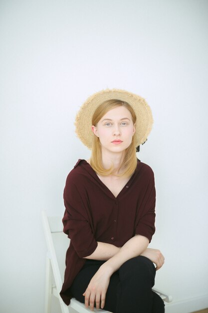 Gorgeous woman sitting and looking in brown shirt, black pants and sun hat in room with white background.