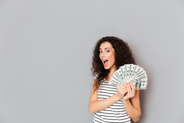 Gorgeous woman in striped t-shirt holding fan of 100 dollar bills in hands smiling on camera being happy and lucky over grey wall