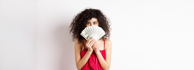 Gorgeous woman with curly hair hiding face behind dollars and smiling standing over white background