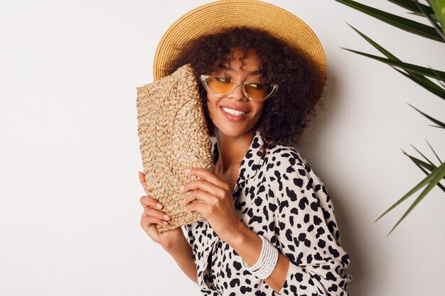 Gorgeous woman with dark skin  in jeans and straw hat posing in studio over white background with  bag in Bali style. Sopping  mood.