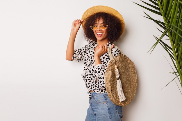 Free photo gorgeous woman with dark skin  in jeans and straw hat posing in studio over white background with  bag in bali style. sopping  mood.