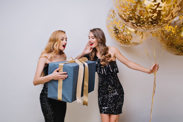 Gorgeous woman with elegant hairstyle holding big gift with surprised face expression. Indoor photo of two pretty girls having fun during celebration and posing