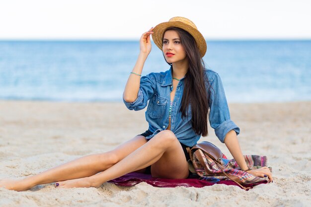Gorgeous woman with tan body, full red lips and l long legs posing on the tropical sunny beach. Wearing crop top, shorts and straw hat.