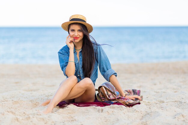 Gorgeous woman with tan body, full red lips and l long legs posing on the tropical sunny beach. Wearing crop top, shorts and straw hat.