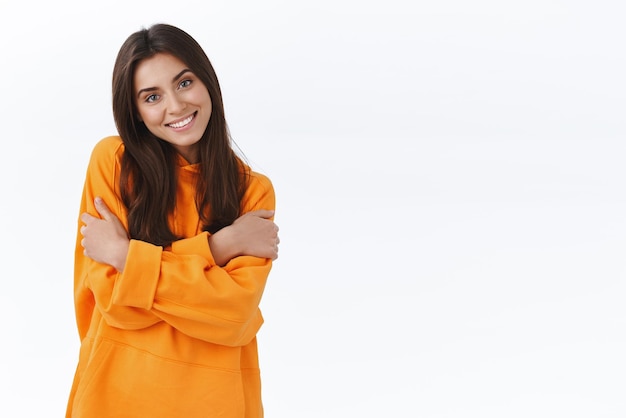 Gorgeous young brunette female in orange hoodie embrace herself cuddle and smile camera tilt head felt chilly walking spring evening borrowed boyfriend hoodie stand white background