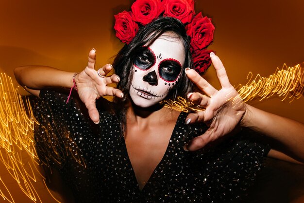 Gorgeous young female model with white face painting posing in halloween Studio photo of debonair zombie girl standing on sparkle background