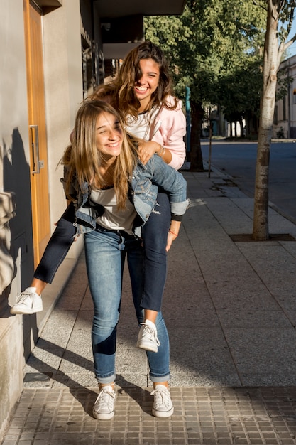 Gorgeous young girl giving piggyback to her female friend on sidewalk