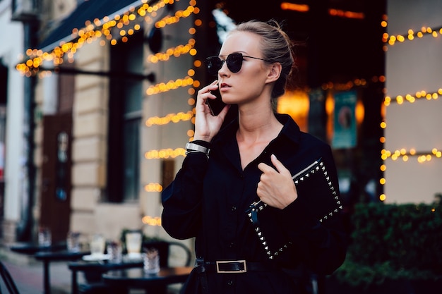 Giovane donna splendida in rigoroso cappotto nero parlando al cellulare, con gli occhiali