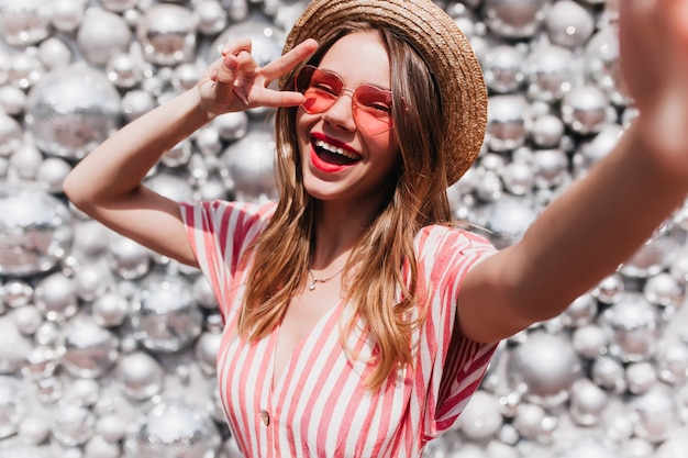 Gorgeous young lady in trendy sunglasses making selfie with disco balls. Fashionable smiling girl in striped attire preparing for summer party.