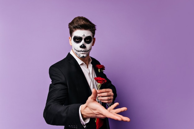 Gorgeous young man with face art in form of skull holding red rose in his hands, posing for portrait on isolated background.