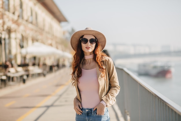 Gorgeous young woman in dark sunglasses standing in confident pose at embankment