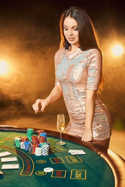 Великолепная молодая женщина в вечернем платье с фишками в руке стоит возле покерного стола с бокалом шампанского
