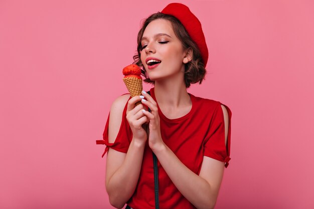 アイスクリームを食べる赤い服のゴージャスな若い女性。デザートでポーズをとる洗練されたフランスの女性モデル。
