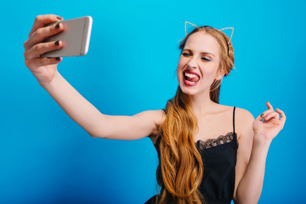 Gorgeous young woman taking selfie, making funny facial expression, showing tongue, at party. She has long blonde hair, nice makeup. Wearing black dress, diadem with cat ears.