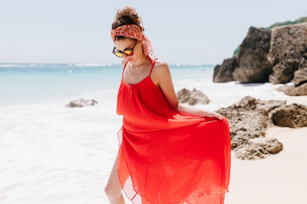 Великолепная молодая женщина с лентой в волосах позирует на берегу моря. Наружное фото романтичной дамы, играющей с длинным красным платьем.