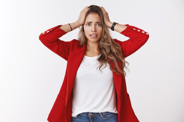 Gosh have trouble. Portrait of panicking worried young cute silly female trainee wearning red jacket holding hands head distressed nervous widen eyes perplexed, having problem white wall