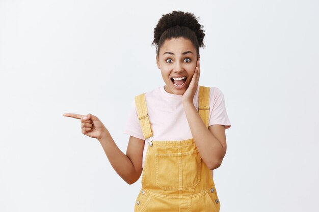 Gosh, what great news. Portrait of surprised good-looking amazed female in yellow stylish dungarees, holding palm on cheek, being charmed and impressed, pointing left and asking question