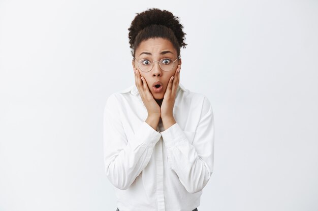 Gosh, what happened to your hair. Portrait of shocked and stunned beautiful dark-skinned female friend with curly hair and glasses, folding lips and staring amazed, holding palms on cheeks