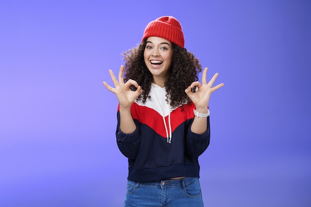 Got under control. portrait of happy charming smiling curly-haired female in warm winter hat and sweatshirt smiling broadly and showing okay or excellent gesture as approving, liking cool movements