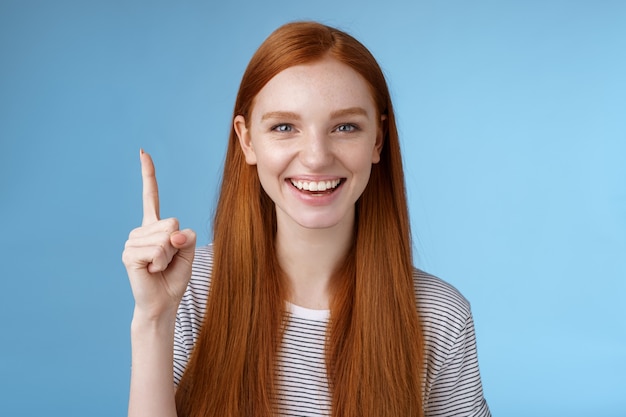 Got excellent idea pick for you. Attractive happy friendly-looking helpful redhead female shop assistant showing gorgeous dress customer pointing up index finger smiling glad help, blue background