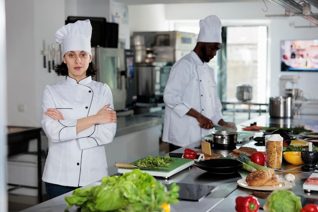 Free photo gourmet cuisine expert standing in restaurant professional kitchen with arms crossed while smiling at camera. confident woman cook with culinary skills preparing ingredients for dinner service.