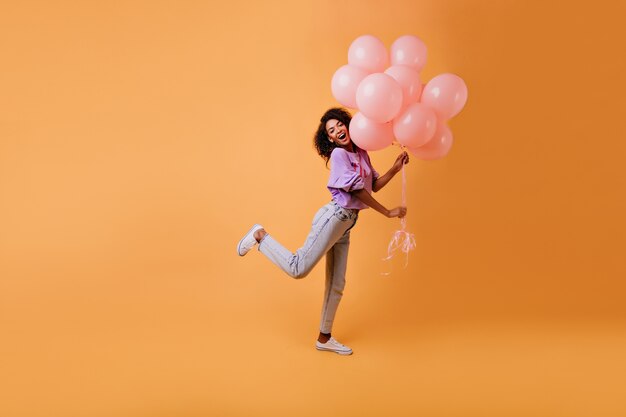 Graceful african female model in casual clothes fooling around on yellow. emotional birthday girl dancing with party balloons.