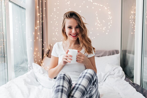 Graceful blissful girl drinking tea with smile. Indoor portrait of dreamy european lady enjoying weekend at home.